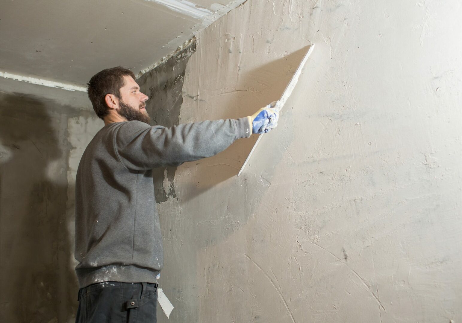 A male plasterer with a beard plasters a concrete wall with a spatula.