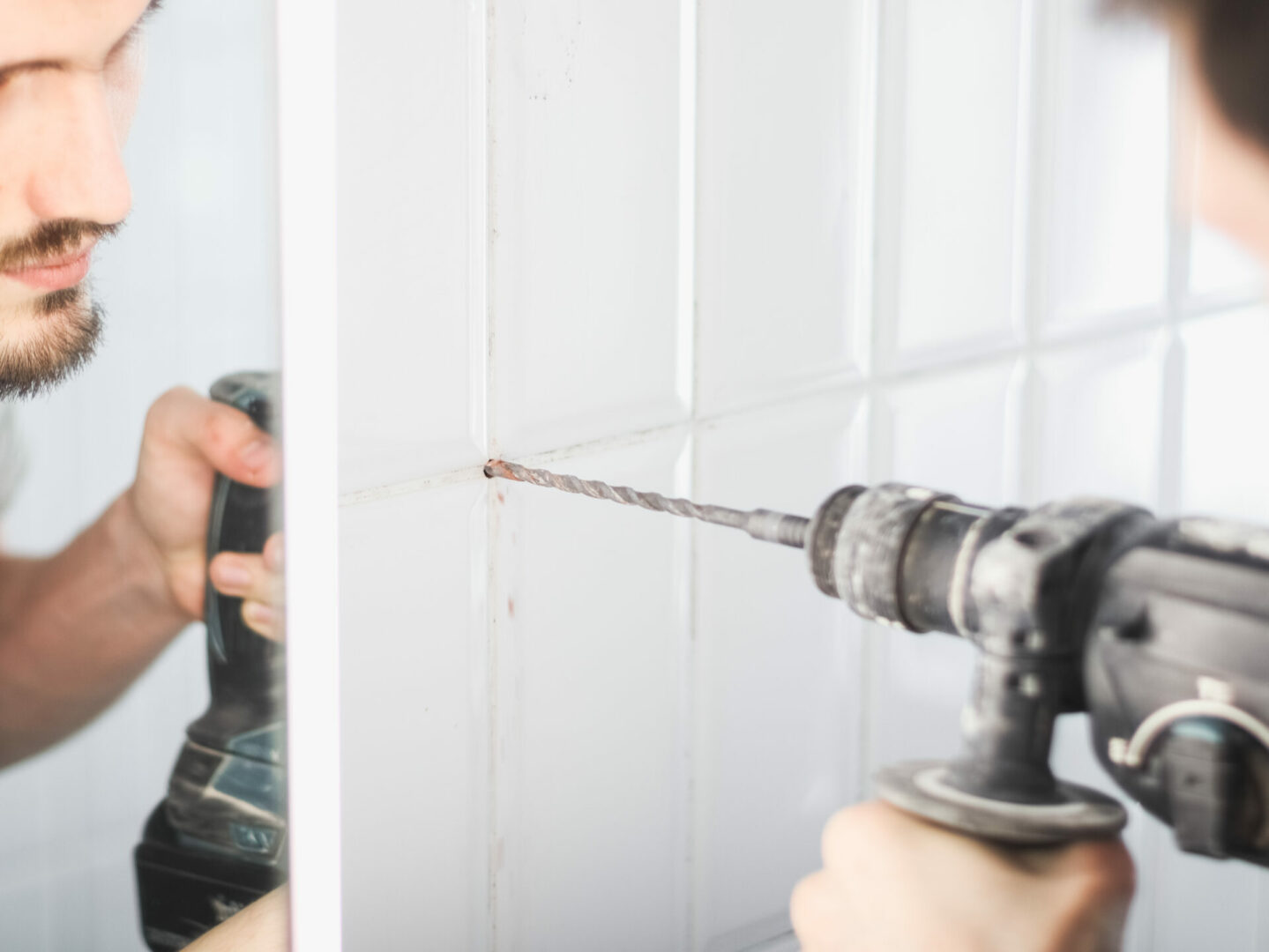 A young man drills between the seams on a tiled wall.